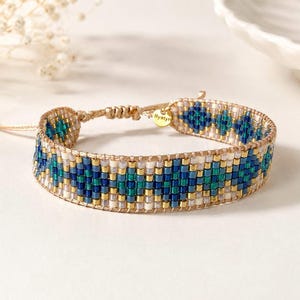 Op de afbeelding: Een geweven kralenarmband met een geometrisch patroon in tinten blauw, groenblauw, wit en metallic goud. De armband heeft een verstelbare koordsluiting en een kleine ronde goudkleurige metalen bedel met de inscriptie Bystyr.