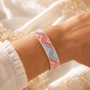Könnte beinhalten: Ein gewebtes Perlenarmband mit einem geometrischen Muster aus kleinen quadratischen Perlen in den Farben Hellblau, Korallenrosa und Metallic-Gold. Das Armband hat eine flache, bandartige Struktur mit einem sich wiederholenden Zickzackmuster.