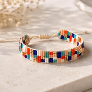 Op de afbeelding: Een geweven armband met een herhalend geometrisch patroon van kleine vierkante kralen in levendige tinten oranje, blauw, turquoise, wit en metallic goud. De armband heeft een verstelbare schuifknoopsluiting gemaakt van dun crèmekleurig koord.