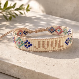 Op de afbeelding: Een geweven armband met een crèmekleurige basis en de tekst "JULY" in beige letters. Het ontwerp bevat geometrische patronen met blauwe, roze en gouden kralen. Het heeft een verstelbare schuifknoopsluiting gemaakt van bijpassend beige koord.