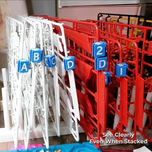 Puede incluir: Piezas de maquetas de plástico blanco y rojo, organizadas en estantes. Cubos azules con letras y números se utilizan para etiquetar. El texto "See Clearly Even When Stacked" es visible. La imagen muestra una configuración de hobby de modelismo.