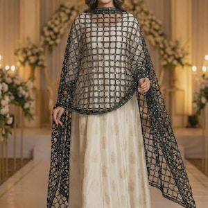 May include: A floor-length cream-colored dress featuring a subtle gold-toned pattern, paired with a black sheer shawl. The shawl has a prominent grid-like lattice design with small reflective embellishments at each intersection of the black fabric.