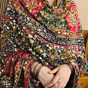 May include: A vibrant shawl featuring a complex pattern of pink, green, yellow, and navy blue fabric. The textile is heavily embellished with small reflective mirrors, gold-toned sequins, and white beads arranged in intricate geometric and floral motifs.
