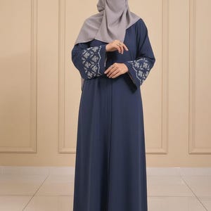Può includere: Abaya blu navy con maniche ricamate e hijab abbinato. L'abito a maniche lunghe presenta ricami bianchi intricati sui polsini. L'hijab è grigio chiaro e l'abaya è lunga fino al pavimento.