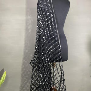 May include: A black, net-like shawl with a grid pattern and sparkling embellishments. The shawl is draped over a black mannequin, showcasing its design and texture. The edges are lined with a row of reflective accents.