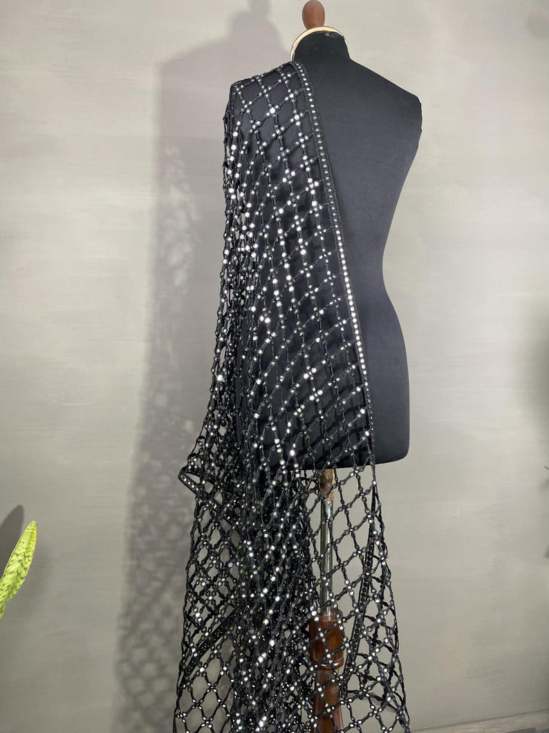 May include: A black, net-like shawl with a grid pattern and sparkling embellishments. The shawl is draped over a black mannequin, showcasing its design and texture. The edges are lined with a row of reflective accents.