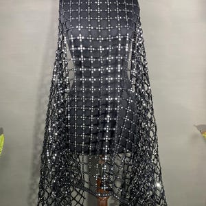 May include: A black garment with a square pattern of silver sequins is displayed on a mannequin. The garment has a net-like overlay that drapes over the shoulders. The mannequin is on a wooden stand against a gray background.