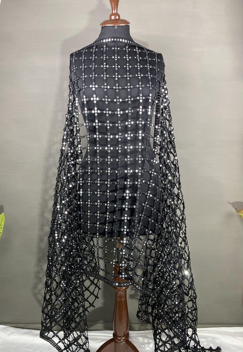 May include: A black garment with a square pattern of silver sequins is displayed on a mannequin. The garment has a net-like overlay that drapes over the shoulders. The mannequin is on a wooden stand against a gray background.
