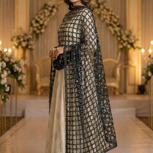 May include: A formal cream-colored dress with a black, grid-patterned overlay. The overlay features sparkling embellishments and long sleeves with fringe. The dress is suitable for special occasions and events.