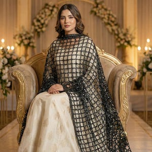 May include: A woman wearing a cream-colored dress with gold floral patterns, and a black, sequined, grid-patterned shawl. She is seated on a gold-colored ornate chair. The background features floral arrangements and soft lighting.
