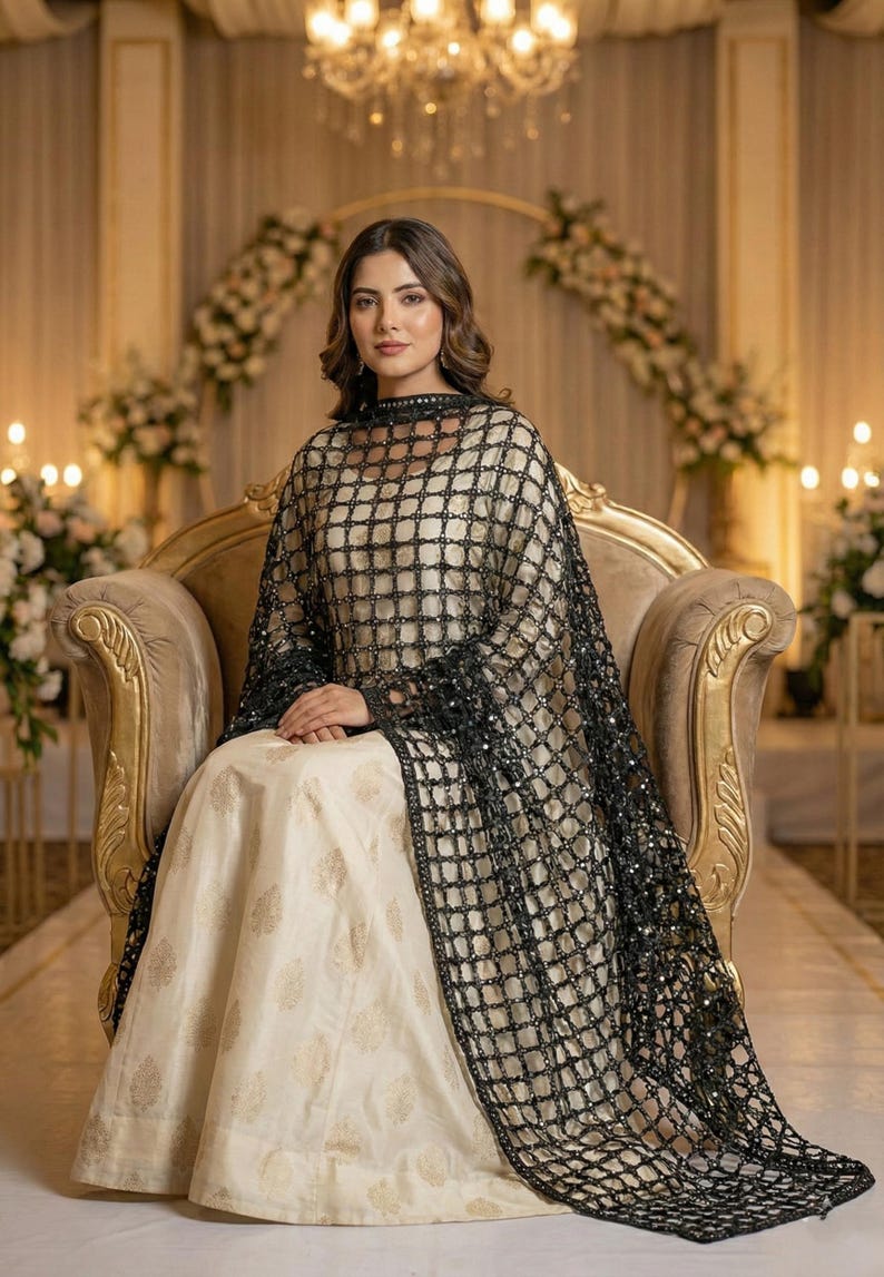 May include: A woman wearing a cream-colored dress with gold floral patterns, and a black, sequined, grid-patterned shawl. She is seated on a gold-colored ornate chair. The background features floral arrangements and soft lighting.