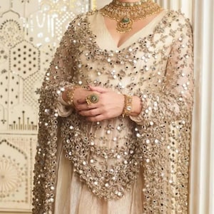 Puede incluir: Una mujer con un vestido color crema y una capa transparente adornada con pequeños adornos reflectantes. Lleva un collar, pulsera y anillo dorados. El fondo presenta una pared decorativa. El conjunto es elegante y ornamentado.