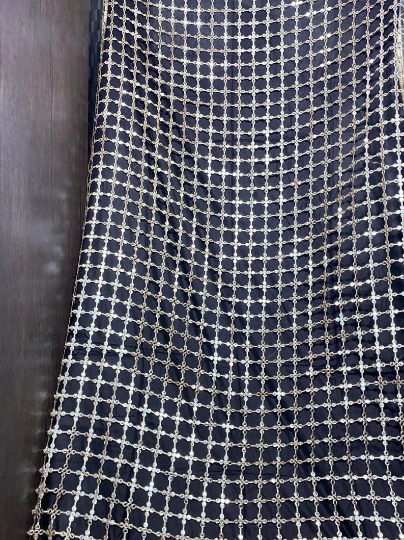 May include: Black fabric with a grid pattern of silver-colored, circular embellishments. The fabric is draped, creating folds and shadows. The overall effect is one of luxury and texture, suitable for evening wear or decorative purposes.