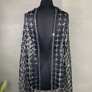 May include: A black, open-weave cape with a grid pattern, adorned with numerous small, reflective embellishments. The cape is draped over a black mannequin, showcasing its design and texture. The edges are lined with a row of the same reflective elements.
