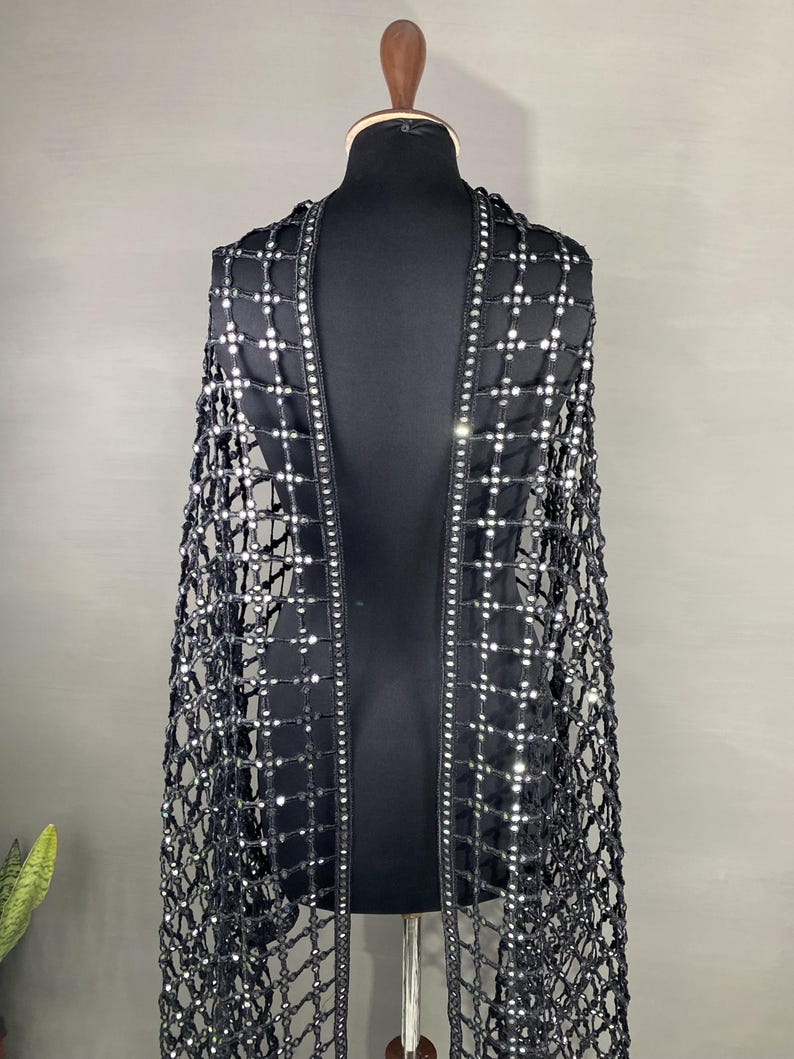 May include: A black, open-weave cape with a grid pattern, adorned with numerous small, reflective embellishments. The cape is draped over a black mannequin, showcasing its design and texture. The edges are lined with a row of the same reflective elements.