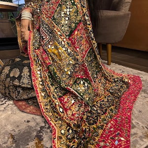 Könnte beinhalten: Ein farbenfrohes, gemustertes Textil mit einer lebendigen Mischung aus Rot, Grün, Gelb und Schwarz. Der Stoff ist mit kleinen, reflektierenden Verzierungen versehen. Die Kanten sind mit einem leuchtend rosa Rand versehen. Das Textil scheint über einer Person drapiert zu sein.