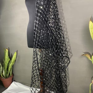 May include: A black, open-weave shawl with a grid pattern, adorned with sparkling embellishments. The shawl is draped over a black mannequin, showcasing its design. The fabric appears to be lightweight, suitable for layering.