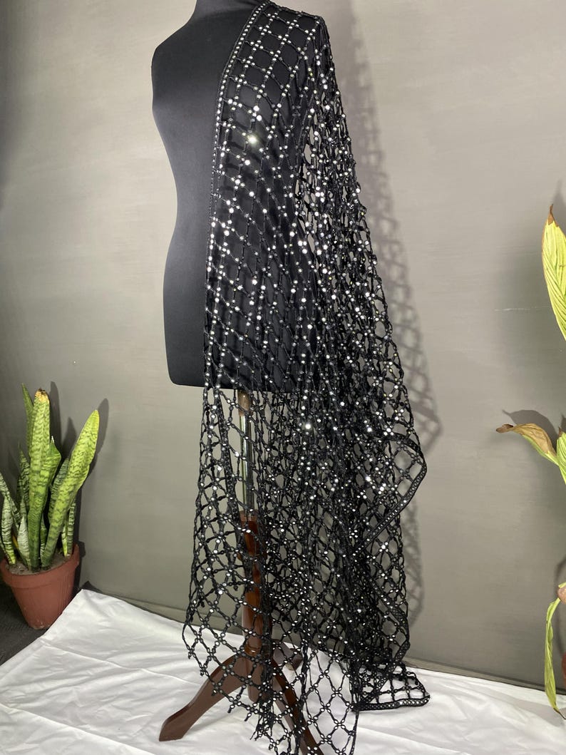 May include: A black, open-weave shawl with a grid pattern, adorned with sparkling embellishments. The shawl is draped over a black mannequin, showcasing its design. The fabric appears to be lightweight, suitable for layering.