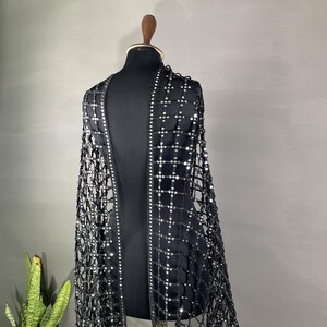 May include: A black, open-front garment with a net-like design, adorned with small, reflective embellishments. The garment is displayed on a black mannequin, showcasing its intricate pattern and draping. The design features a grid-like structure with sparkling accents.