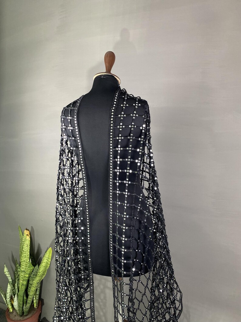 May include: A black, open-front garment with a net-like design, adorned with small, reflective embellishments. The garment is displayed on a black mannequin, showcasing its intricate pattern and draping. The design features a grid-like structure with sparkling accents.