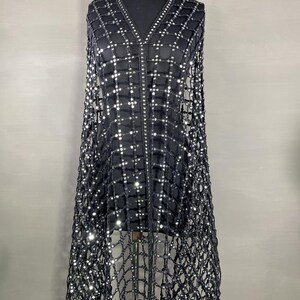 May include: A black, open-weave cape with a grid pattern, embellished with small, reflective sequins. The cape features a V-neck and a central zipper. The garment is displayed on a mannequin against a neutral background, showcasing its draping design.