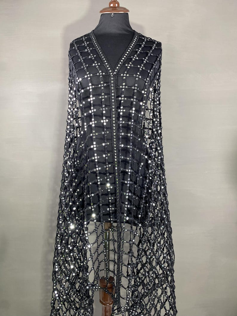 May include: A black, open-weave cape with a grid pattern, embellished with small, reflective sequins. The cape features a V-neck and a central zipper. The garment is displayed on a mannequin against a neutral background, showcasing its draping design.