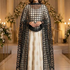 May include: A formal cream-colored gown with gold floral patterns, paired with a black, sequined, grid-patterned cape. The cape has long sleeves and a matching trim. The outfit is suitable for special occasions.
