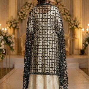 May include: A formal cream-colored dress with a gold pattern is worn under a black, square-patterned, beaded shawl. The shawl drapes over the shoulders. The background features floral arrangements and soft lighting.