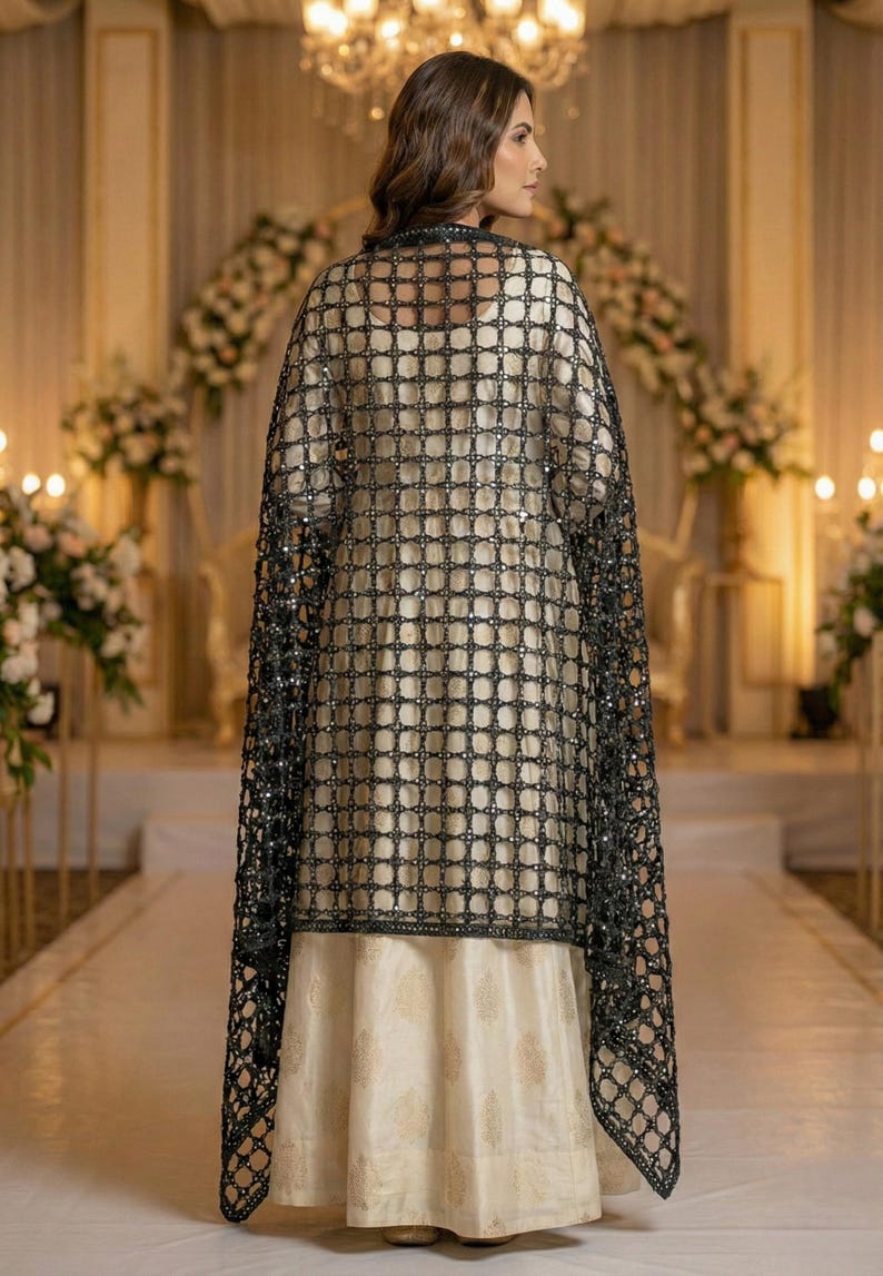 May include: A formal cream-colored dress with a gold pattern is worn under a black, square-patterned, beaded shawl. The shawl drapes over the shoulders. The background features floral arrangements and soft lighting.