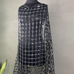 May include: A black, open-weave shawl with a grid pattern of small, reflective embellishments. The shawl is draped over a black mannequin, showcasing its design and texture. The garment has a V-neckline and a fringed edge.