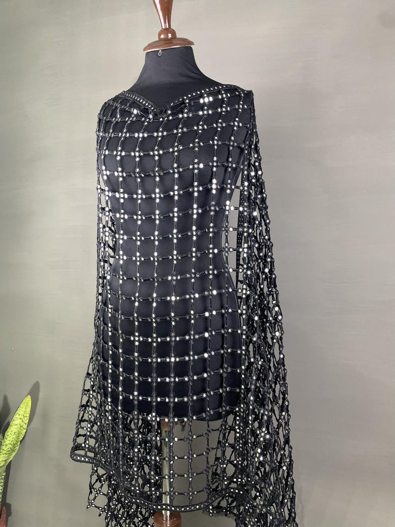 May include: A black, open-weave shawl with a grid pattern of small, reflective embellishments. The shawl is draped over a black mannequin, showcasing its design and texture. The garment has a V-neckline and a fringed edge.