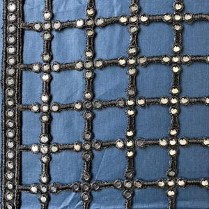 May include: Close-up of a blue fabric with a grid-like pattern. The grid is formed by black, woven material, and each intersection features a small, round, reflective mirror. The edges are trimmed with a similar black, beaded border.