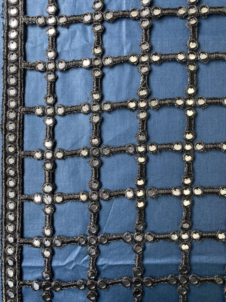 May include: Close-up of a blue fabric with a grid-like pattern. The grid is formed by black, woven material, and each intersection features a small, round, reflective mirror. The edges are trimmed with a similar black, beaded border.
