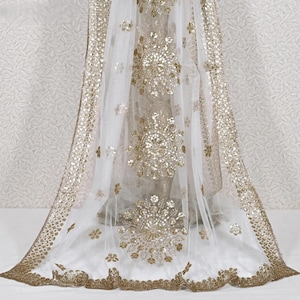 May include: A white sheer fabric with gold embroidery and embellishments. The design features large, circular floral patterns and a detailed border. The fabric is draped, showcasing the delicate texture and shimmering details.