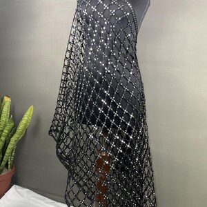May include: A black, open-weave shawl with a grid pattern, adorned with reflective embellishments. The shawl is draped over a mannequin, showcasing its design and texture. The fabric appears to be lightweight and suitable for layering.