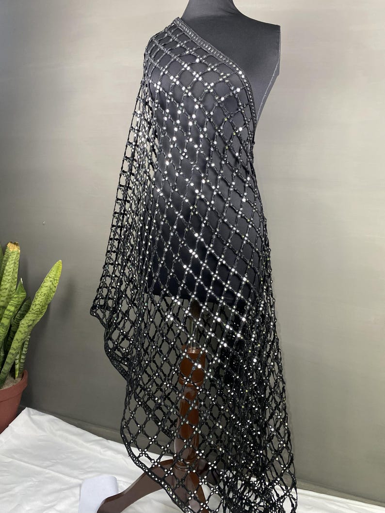 May include: A black, open-weave shawl with a grid pattern, adorned with reflective embellishments. The shawl is draped over a mannequin, showcasing its design and texture. The fabric appears to be lightweight and suitable for layering.