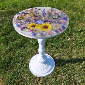 Può includere: Un tavolino bianco con piano rotondo con design floreale in resina. Il tavolo ha una base a piedistallo ed è appoggiato sull'erba. Il piano del tavolo è intarsiato con fiori secchi colorati, tra cui girasoli e altri fiori selvatici.