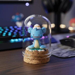 May include: A blue and yellow cartoon character figurine inside a clear glass dome. The figurine is on a cork base. The background includes a keyboard, computer monitor, and headphones.