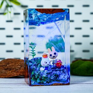 May include: A rectangular resin art piece featuring two cartoon animal figures on a blue base. The figures are a white cat and a pink rabbit, each holding a lantern. The top of the piece has a blue and brown design.
