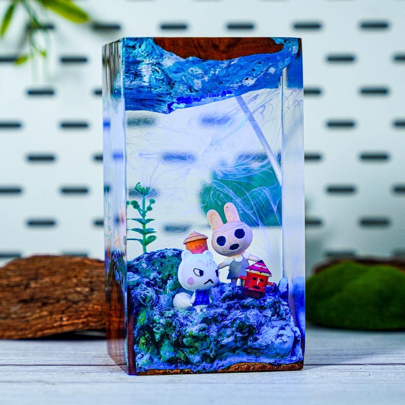 May include: A rectangular resin art piece featuring two cartoon animal figures on a blue base. The figures are a white cat and a pink rabbit, each holding a lantern. The top of the piece has a blue and brown design.