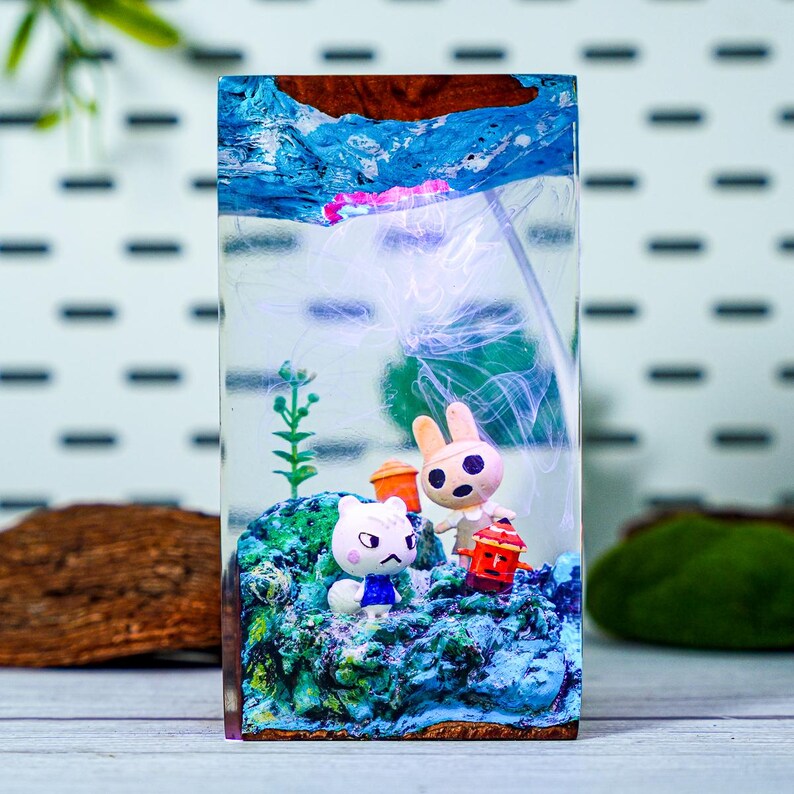 May include: A rectangular resin art piece featuring a miniature underwater scene. The scene includes two cartoon animal figures, a white cat and a rabbit, on a blue and green base. The top of the piece has a blue and brown design, resembling water and land.