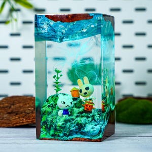 May include: A rectangular resin art piece featuring miniature figurines of cartoon characters on a green, mossy base. The resin has blue and turquoise swirls, with a wooden frame. The figurines include a cat and a rabbit.