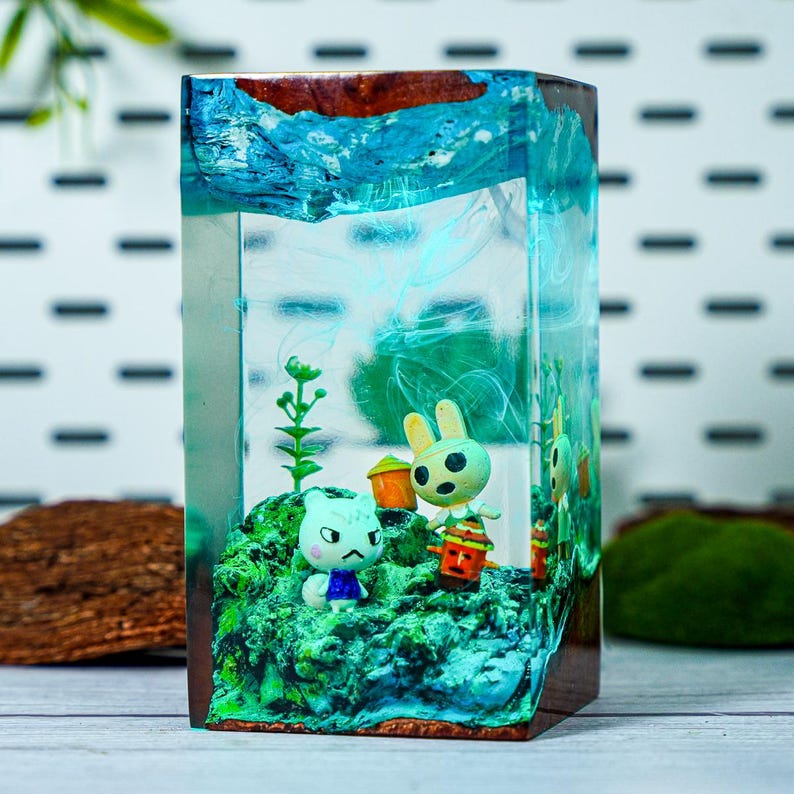 May include: A rectangular resin art piece featuring miniature figurines of cartoon characters on a green, mossy base. The resin has blue and turquoise swirls, with a wooden frame. The figurines include a cat and a rabbit.