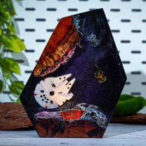 Peut inclure: Une pièce d'art en résine hexagonale représentant une scène spatiale. Elle comprend un Faucon Millenium blanc, des chasseurs stellaires et une structure planétaire avec un cratère rouge. Le fond est un ciel sombre et étoilé.