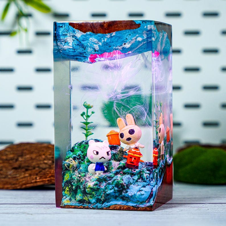 May include: A rectangular resin art piece featuring miniature figurines of cartoon characters in a diorama setting. The scene includes a white cat, a rabbit holding a lantern, and other figures on a blue and green landscape, with a blue and pink sky.