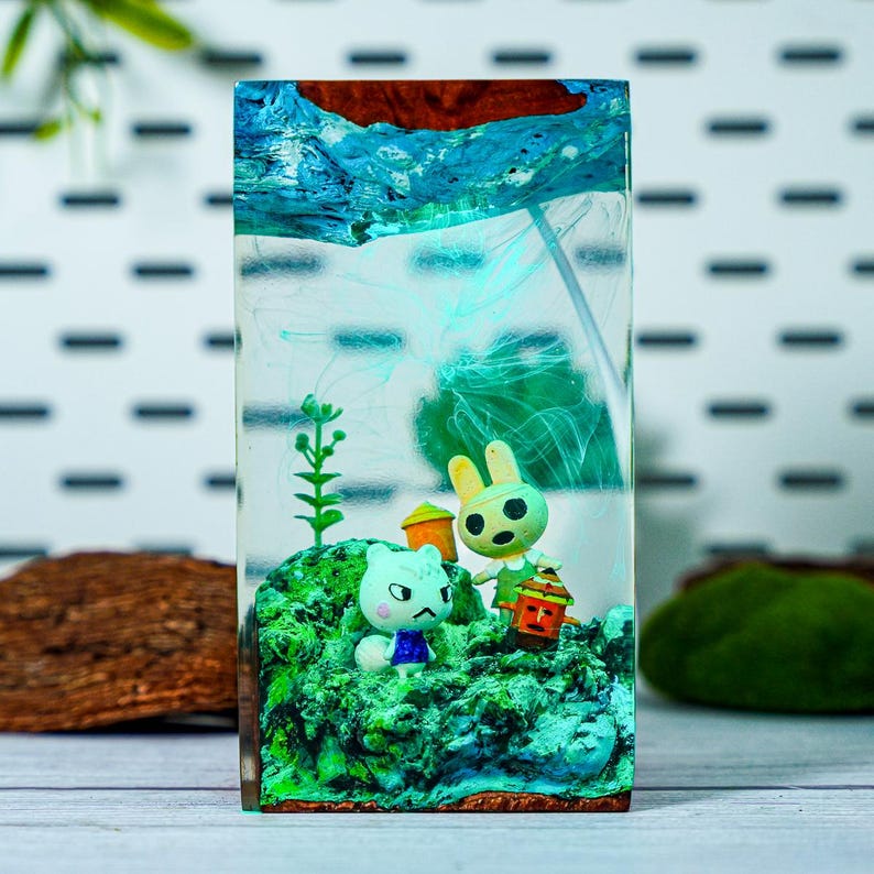 May include: A rectangular resin art piece featuring a miniature diorama. The scene includes two cartoon animal figures, a rabbit and a cat, on a green, mossy base. The resin has blue and green swirls, with a wooden top.