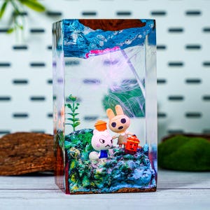 May include: A clear resin block featuring a miniature diorama with two cartoon animal figures, a rabbit and a cat, standing on a colorful, textured base. The diorama includes a small plant and a painted blue and brown background.