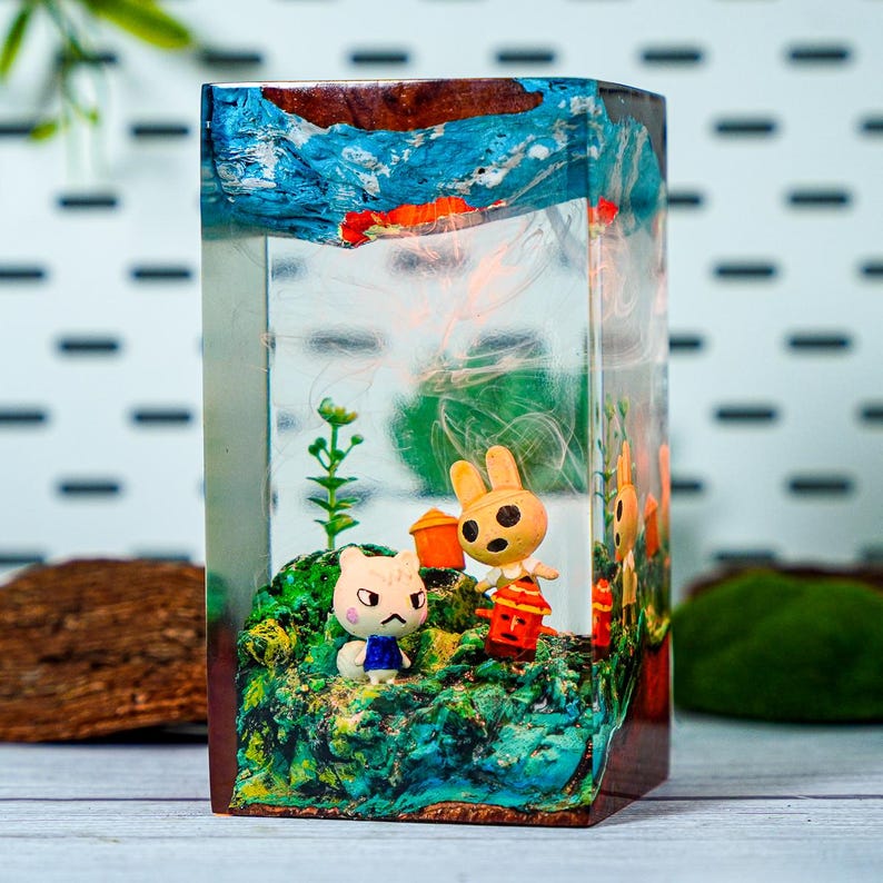 May include: A rectangular resin art piece featuring miniature figurines of cartoon animals on a green and blue landscape. The top has blue and orange abstract art. The piece is encased in clear resin.