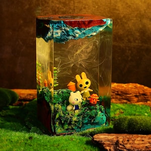 May include: A clear resin block with a miniature diorama inside. The diorama features cartoon characters, green moss, and small orange structures. The top is a mix of blue and brown, resembling a landscape. The overall effect is a whimsical, artistic piece.