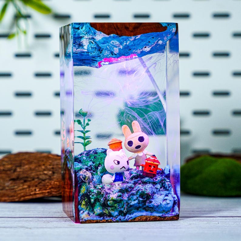May include: A rectangular resin art piece featuring two cartoon animal figures on a rocky base. The clear resin encases a miniature scene with blue, green, and brown elements, and a wooden top. A red lantern is held by one of the figures.